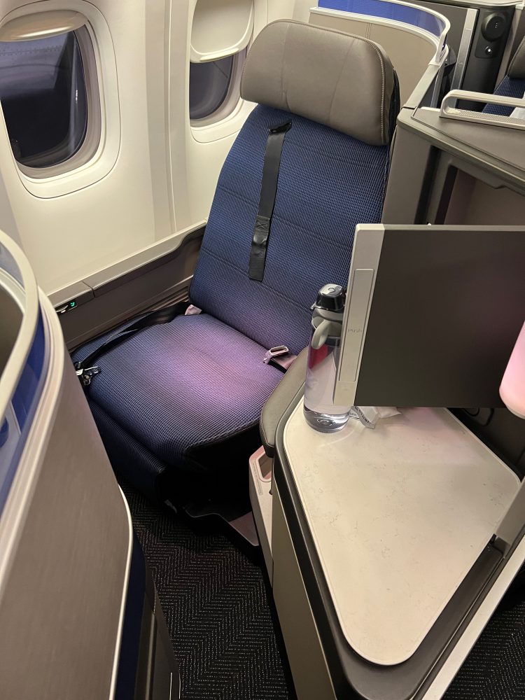 A blue-purple color first class airplane seat with a storage compartment and two windows on the other side of the seat.
