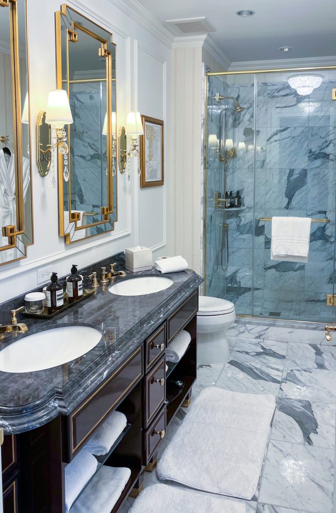 A large white marble lined bathroom with two sinks. There is gold trim on the sinks, mirrors, and shower door.