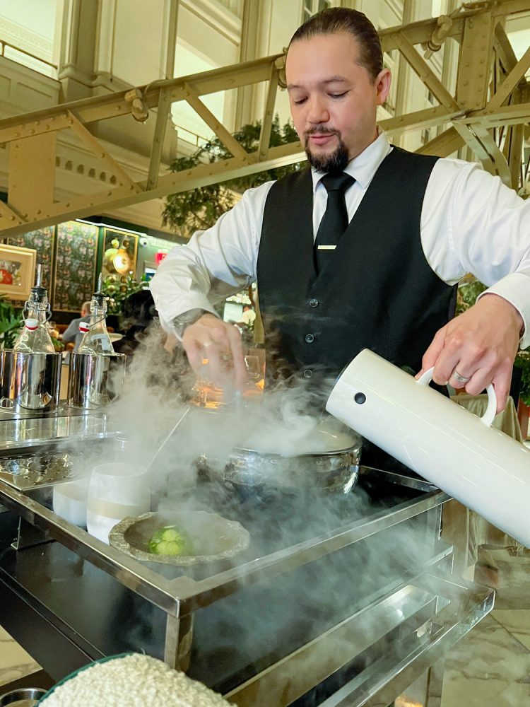 A waiter, in a vest and tie, pours liquid nitrogen from a white container into a bowl while stirring
