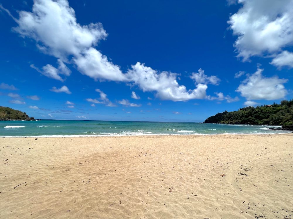 A beautiful white sand beach with clear ocean water beyond the beach. There are small waves. The beach is flanked with land and luscious tropical trees. The sky is mostly clear with a few scattered cumulus clouds.
